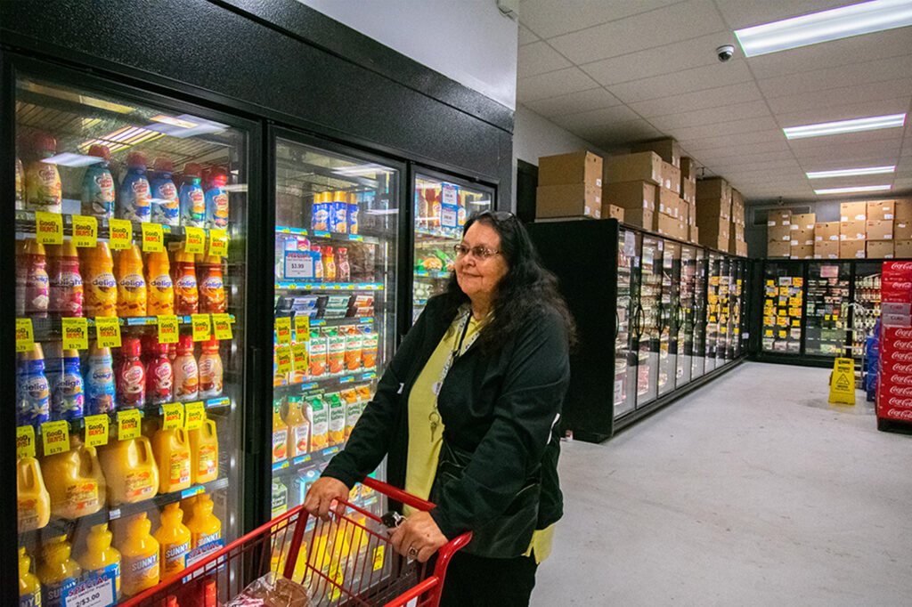 A person shops in a grocery store aisle with refrigerated drinks and boxed goods in a cart. Northern Plains Reservation Aid is relevant.