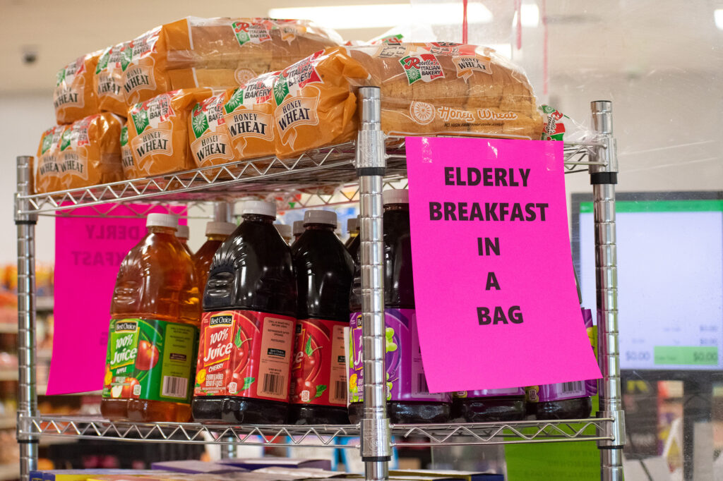 Loaves of honey wheat bread and bottles of juice are displayed with a sign reading "Elderly Breakfast in a Bag."