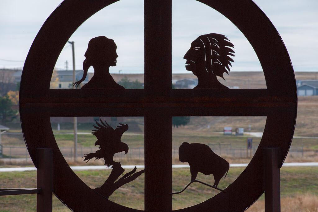 A circular metal sculpture features silhouettes of a woman, a man in a headdress, an eagle, and a bison against a rural backdrop.