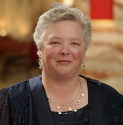 A person in formal attire, wearing earrings and a necklace, stands indoors.
