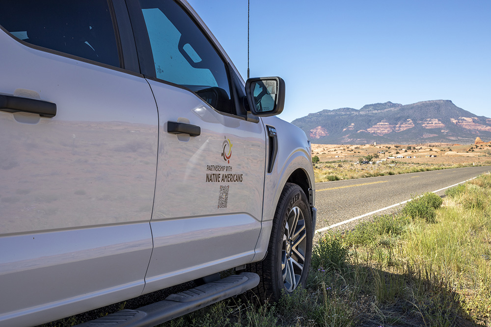 A white truck with the logo "Partnership with Native Americans" parked by a road in a desert landscape with mountains in the distance.