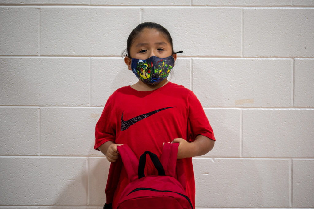 A child wearing a colorful mask and red shirt holds a matching red backpack in front of a white brick wall.