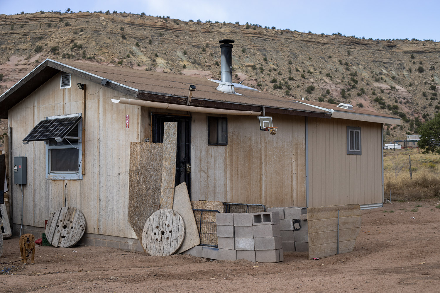 A small, simple house with a solar panel, chimney, cinder blocks, a dog, and desert scenery. Southwest reservation aid is appropriate.