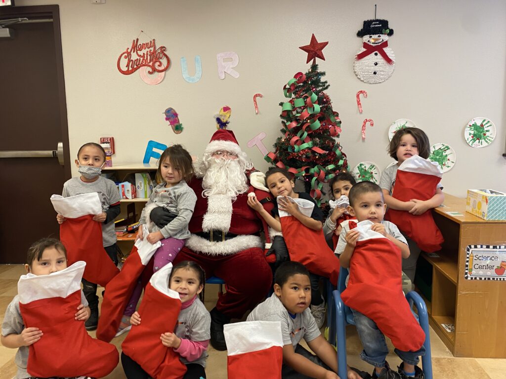 A group of children with Santa, holding stockings, in a festive setting with a Christmas tree and decorations.