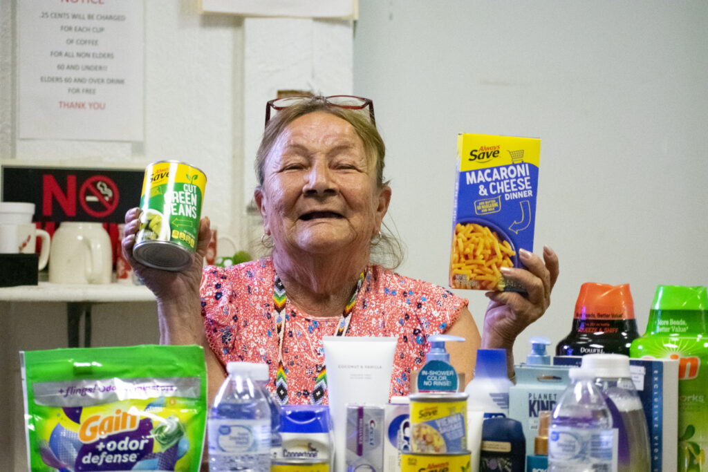 An elderly woman holds a can of green beans and a box of macaroni and cheese, surrounded by various groceries.