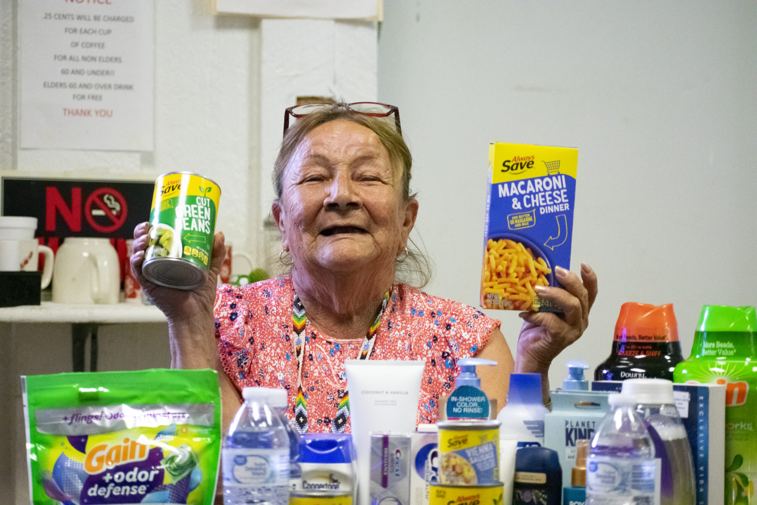 An elderly woman holds a can of green beans and a box of macaroni and cheese, surrounded by various groceries.