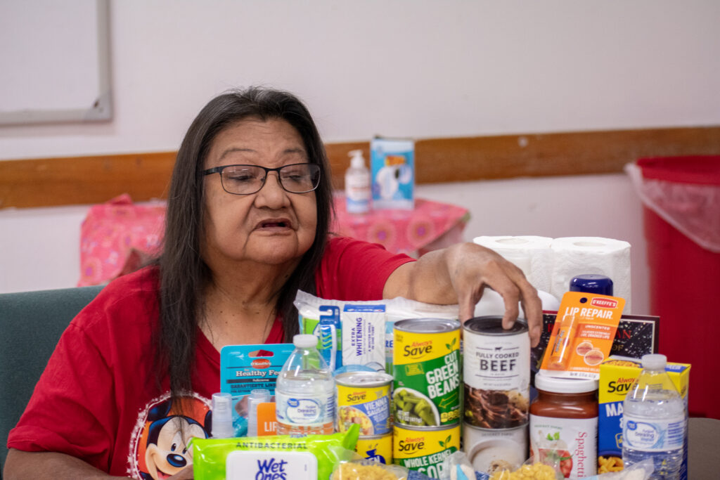 A person is reaching for canned food and supplies on a table. Items include canned beef, vegetables, and toiletries.